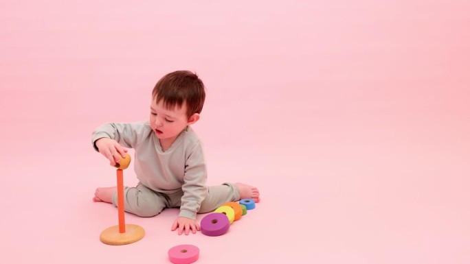 快乐的幼儿在粉红色背景上玩金字塔。小男孩把木环放在玩具金字塔上。孩子一岁八个月，身高正常