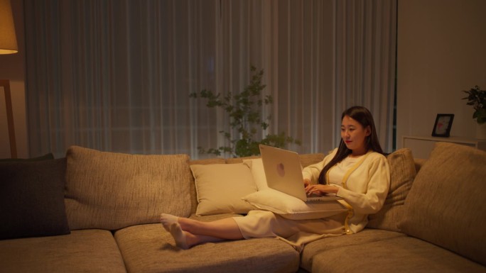 一位韩国女性躺在舒适的沙发上休息，在极简主义的客厅里用笔记本电脑工作到深夜，与一群经理合作进行一个创