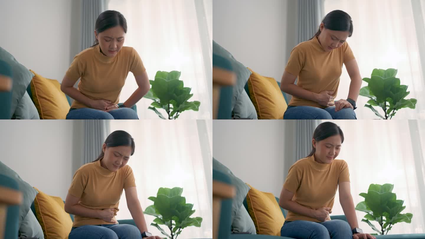 一个亚洲女人肚子疼，坐在家里客厅的沙发上。