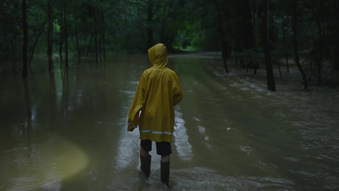 穿着黄色雨衣带着手电筒的男孩在森林里的洪水平原上行走
