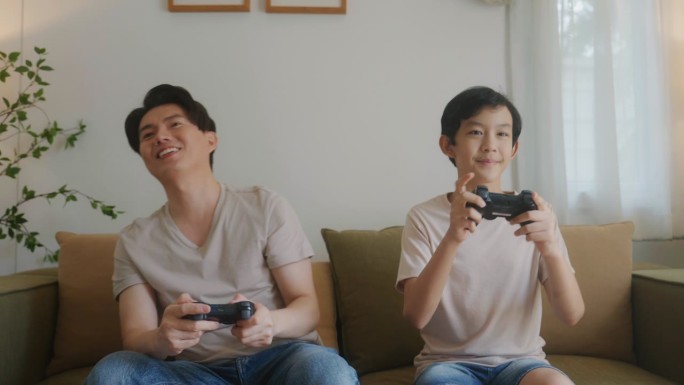 亚洲父亲和孩子在家里用操纵杆玩电子游戏，在度假时一起聊天。