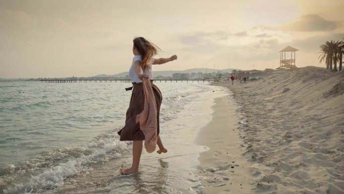 沙滩上夕阳西下的慢动作，一位年轻女子欢快地跳着舞，围着飞舞的丝巾跑着。旅游、度假和冒险的概念