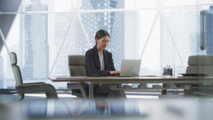 职业女性CEO在明亮的大都市转角办公室用笔记本电脑打字。白人女性独自在投资管理公司的会议室工作，寻找