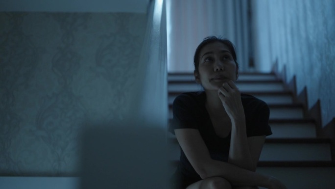 体贴的亚洲妇女坐在楼梯上，手放在下巴上，抬头望向别处，在黑暗的房间里等待家人回家。