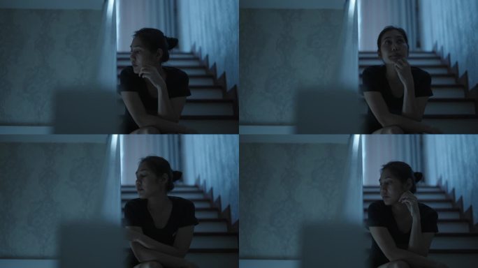 体贴的亚洲妇女坐在楼梯上，手放在下巴上，抬头望向别处，在黑暗的房间里等待家人回家。