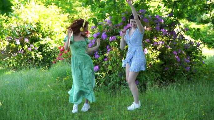 两个快乐的女人的户外肖像，女朋友，朋友穿着裙子戴着耳机在公园里跳舞。随时随地听在线音乐，做你自己的概