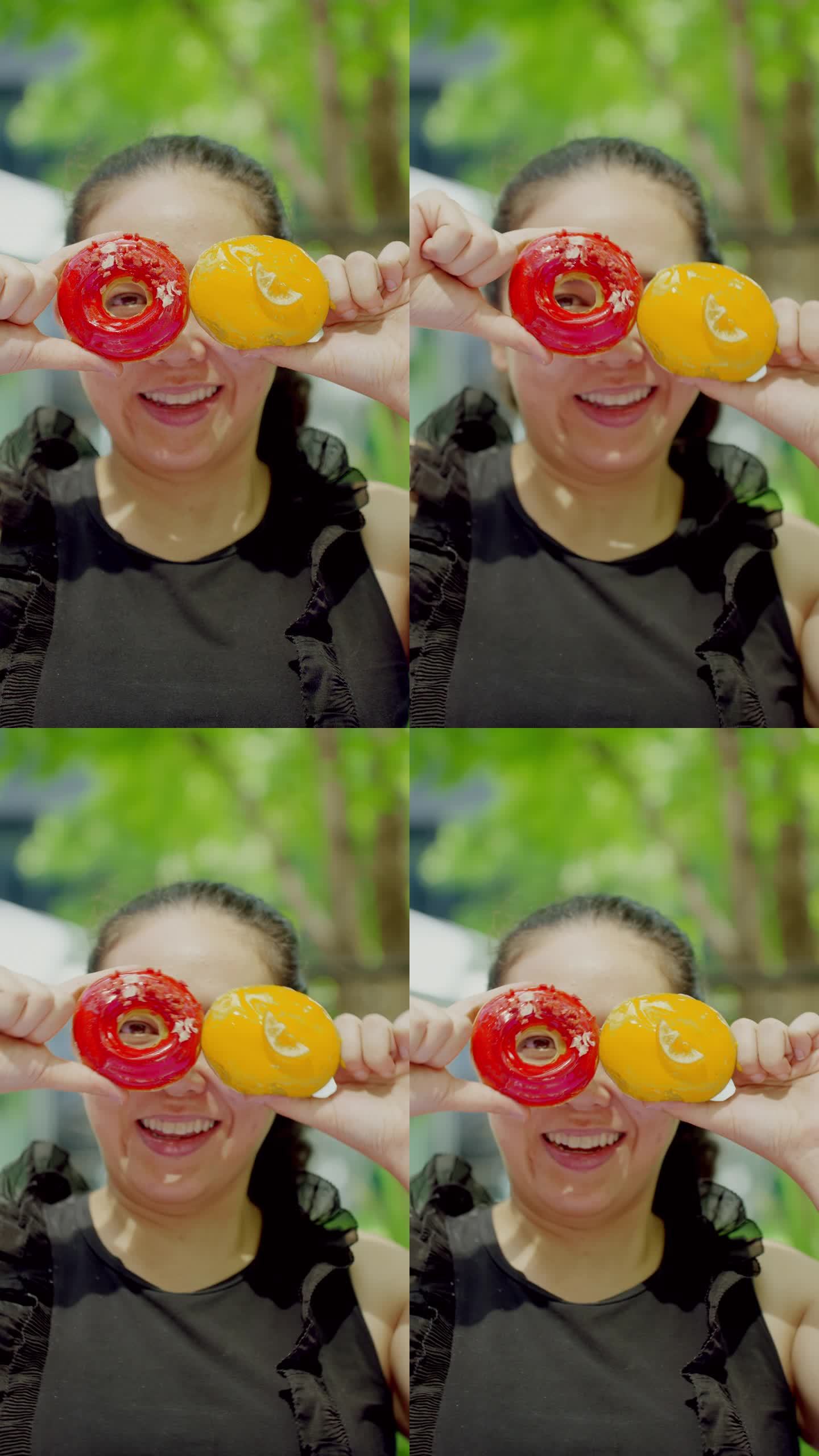 特写:看着镜头:亚洲中年妇女双手拿着两个红色和黄色的甜甜圈，像跳舞的甜甜圈一样和摄影师一起玩耍和移动