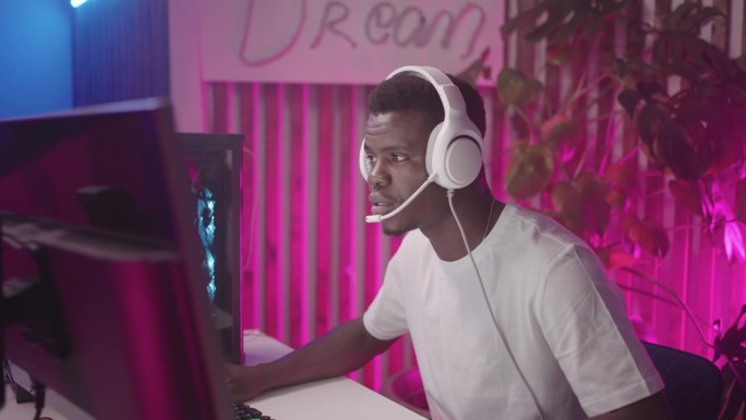 在电脑俱乐部玩电脑游戏的非洲年轻人，戴着耳机的快乐黑人男性肖像，与玩伴交流，青少年的网络和电子游戏成