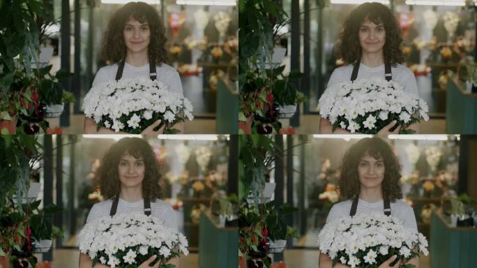 穿着制服的快乐女售货员站在花店里，拿着美丽的花束，看着镜头