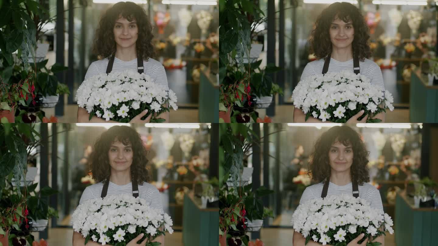 穿着制服的快乐女售货员站在花店里，拿着美丽的花束，看着镜头