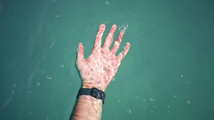 男子将手浸入湛蓝河水的慢动作。男性手与黑色智能手表在手腕上的特写，在清澈透明的水中加深