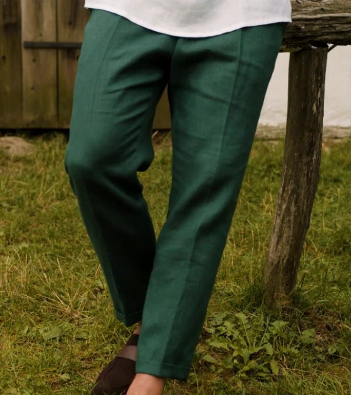 一个穿着白色绣花衬衫和绿色裤子的模特躺在草地上，背景是一座木屋。一个穿着绿裤子和黑鞋的模特的腿。垂直