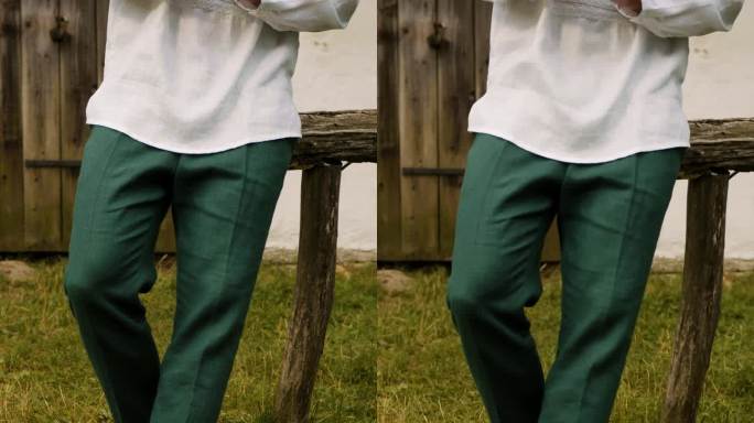 一个穿着白色绣花衬衫和绿色裤子的模特躺在草地上，背景是一座木屋。一个穿着绿裤子和黑鞋的模特的腿。垂直