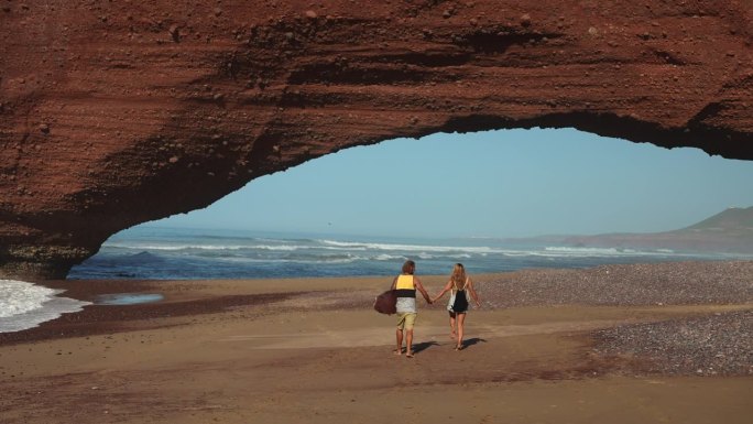 一对情侣手牵手走在桥状岩石下的沙滩上，背景是大海