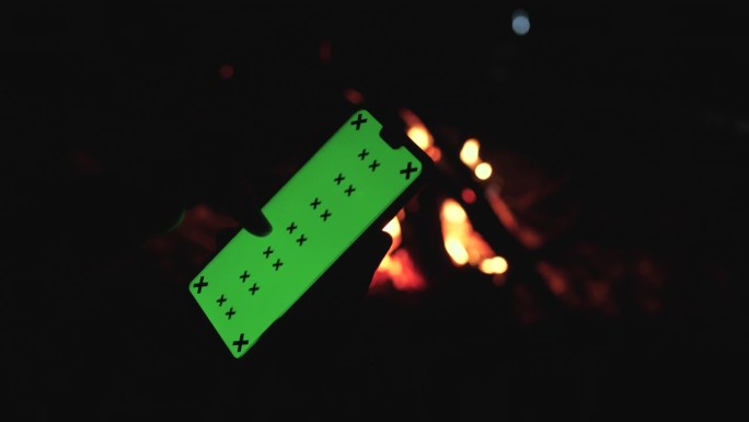男子手持智能手机在肖像模式与绿色屏幕模型旁边的营火在晚上