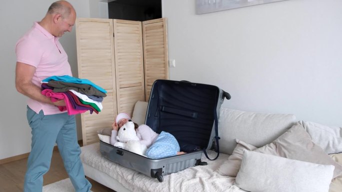 男人用手提箱打包东西。一个7-8岁的白人女孩从一个大行李箱里躲了出来，想要吓唬她的父亲。