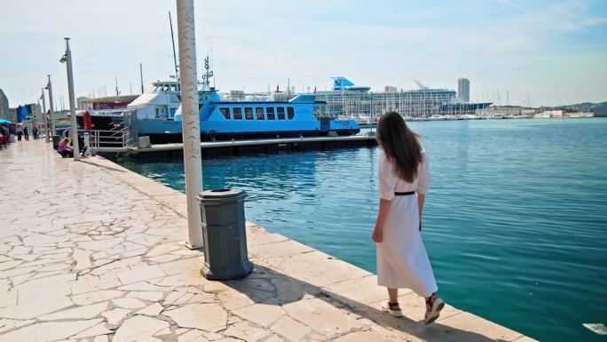 在一个阳光明媚的日子里，一名年轻女子穿着裙子走在法国土伦的克鲁斯港边。