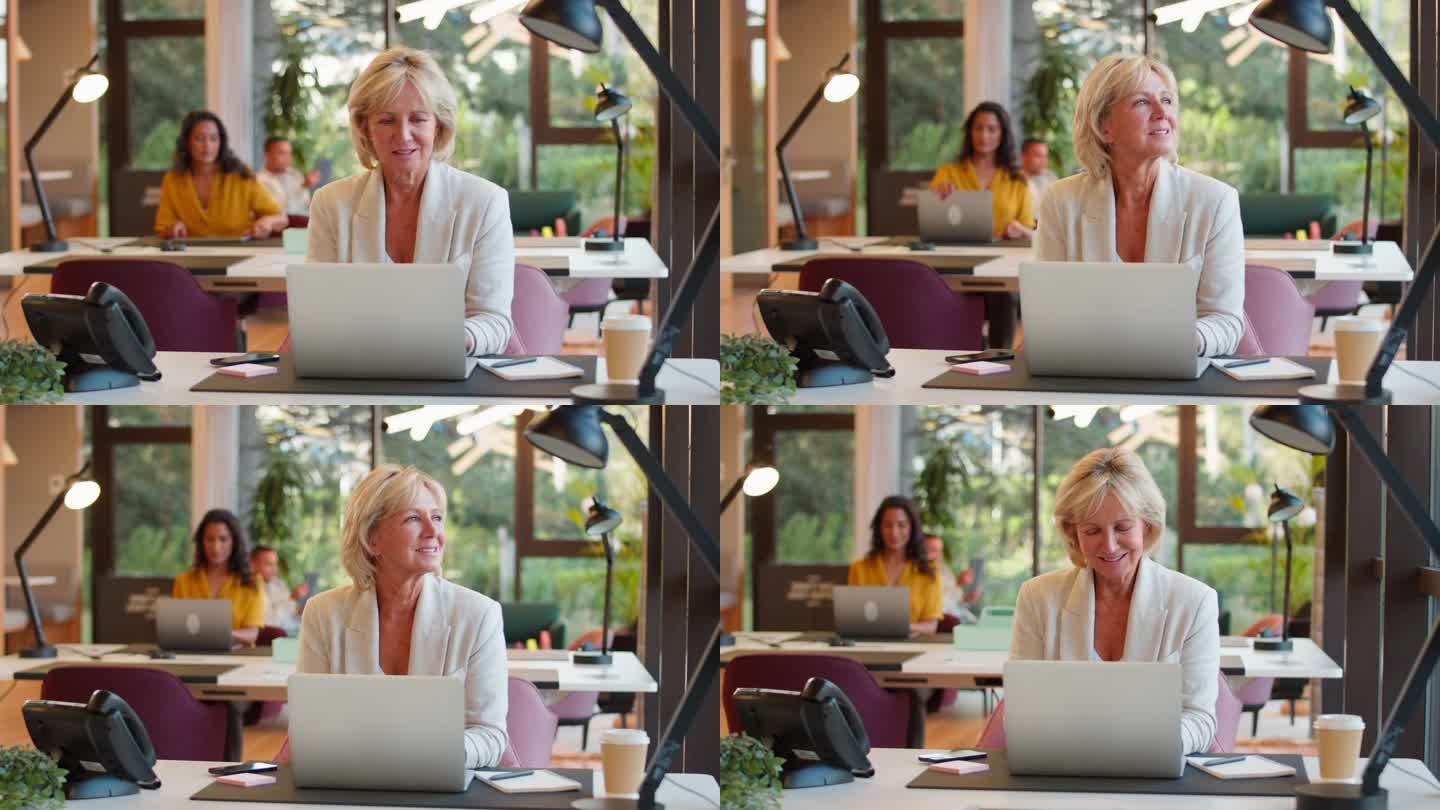 成熟的女商人在办公室的桌子上用笔记本电脑工作，停下来看看窗外