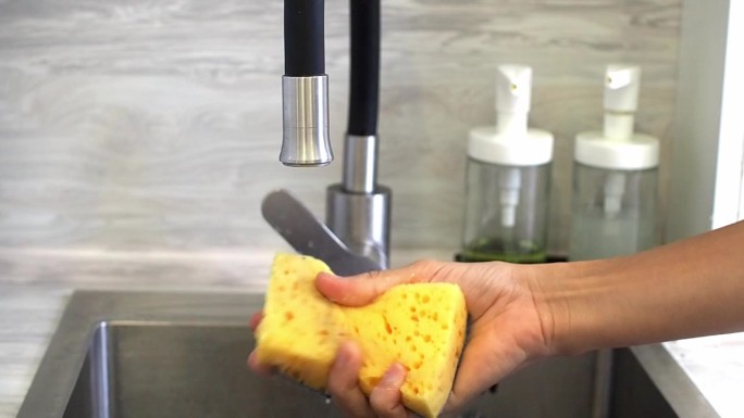 在厨房的灰色水槽里，一个女人用清洁剂压着一块黄色泡沫海绵