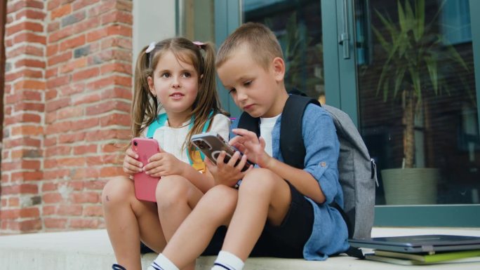 欢快时尚的小学生小男孩小女孩坐在户外楼梯上使用手机上网交流。友谊，科技理念。在线玩游戏。