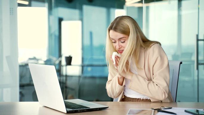 患有季节性过敏症的年轻女商人坐在现代办公室的办公桌前用笔记本电脑工作。