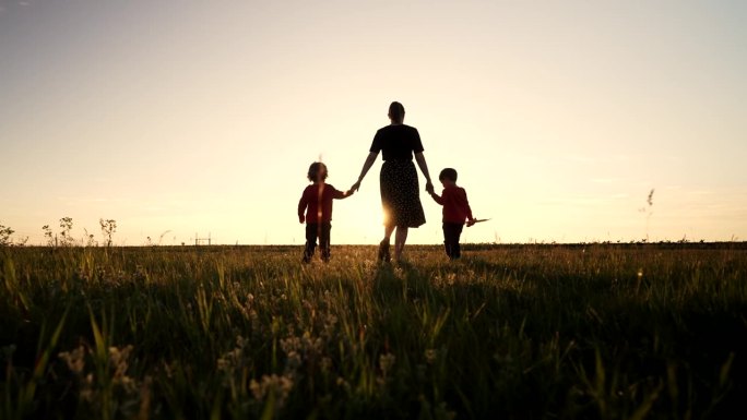 年轻家庭的剪影——母亲和两个兄弟双胞胎男孩一起走在户外或公园的阳光下，黄金时刻，闪耀着快乐的母亲和孩