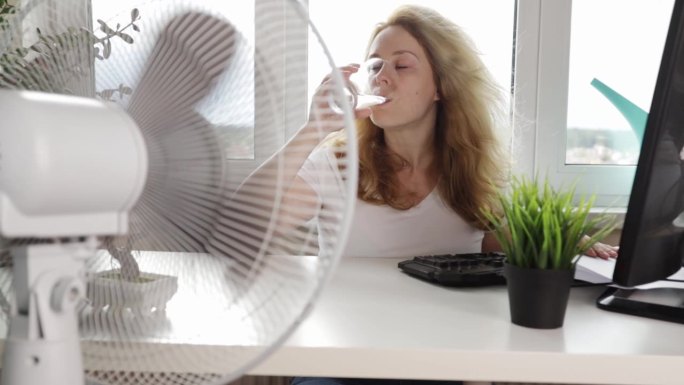 炎热的办公室，女人吹风扇保持凉爽和舒适，试图在一个没有空调的办公室里降温，上班族喝水，脱水，中暑，夏