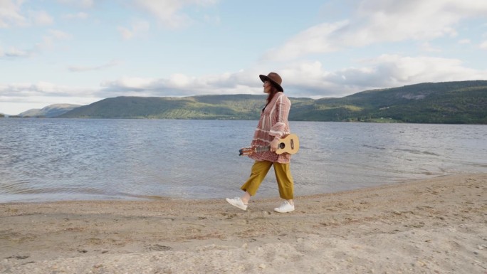 戴着帽子的时髦女人沿着海滩散步。手里拿着吉他，衣服在风中飘动。慢动作