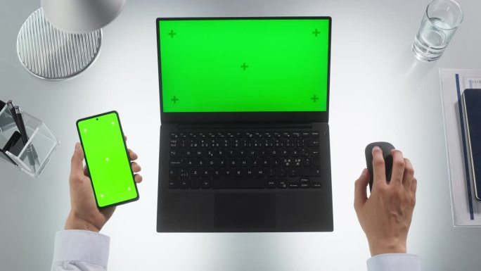 一个匿名的人用笔记本电脑和现代智能手机进行多任务处理的自上而下的视图。这两个设备都有模拟绿屏显示与运