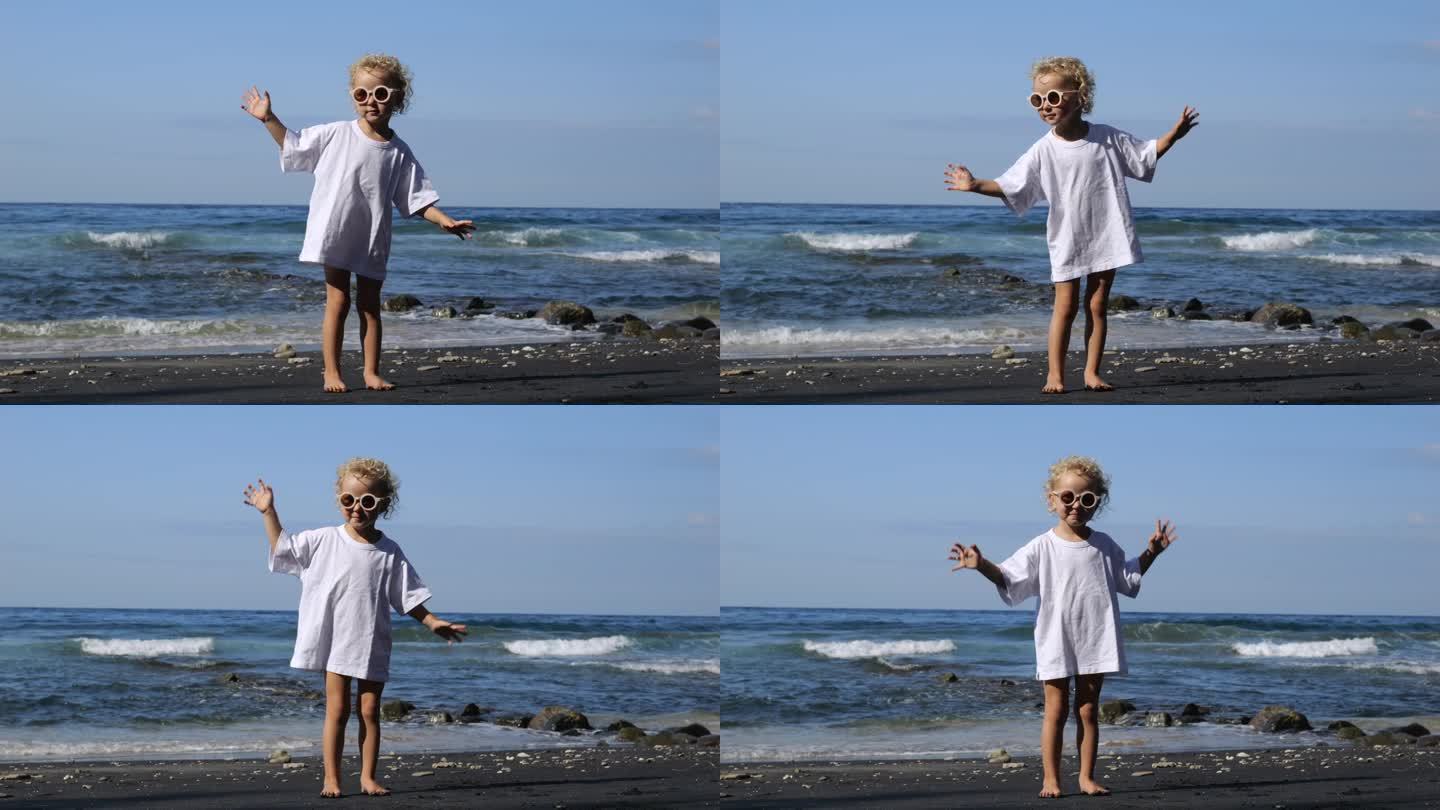 周末，穿着白色t恤的小女孩在海边晒太阳。可爱的宝宝站在岸上跳舞，玩得很开心。周末假期，在天堂般的美丽
