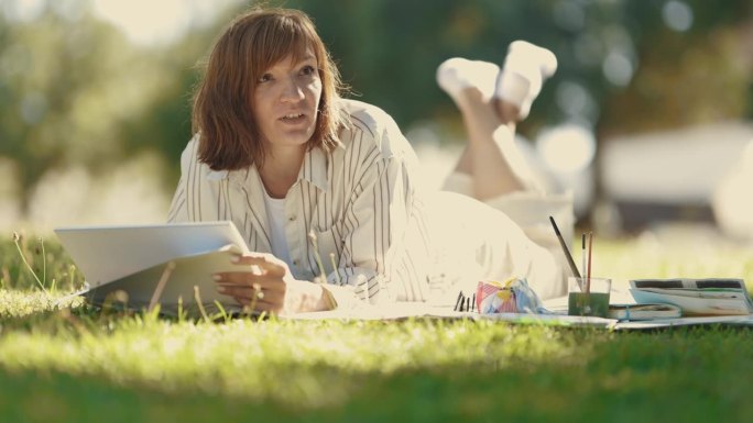一个成年妇女躺在盛开的公园里的毯子上——拿着一本速写本和别人说话