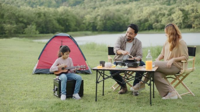 暑假周末，可爱的女儿在露营帐篷里唱歌和弹奏尤克里里琴，与父母在草地上放松，野餐。