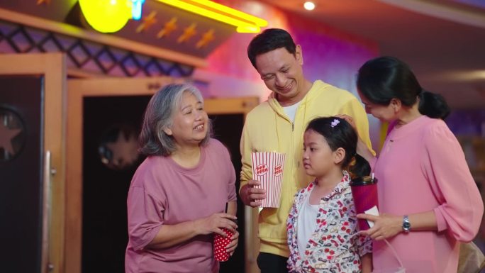 幸福的亚洲家庭喜欢站在电影院前聊天。亚洲家庭拿着碳酸饮料和一桶爆米花一起在电影院等电影。家庭活动