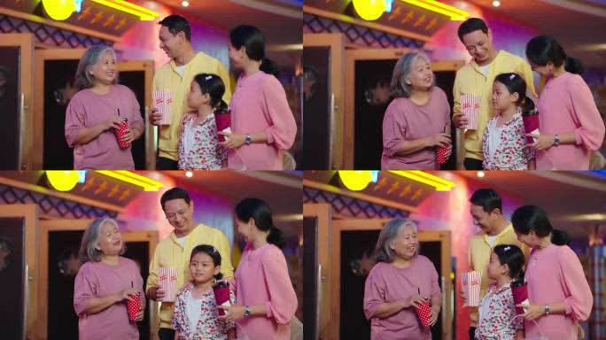 幸福的亚洲家庭喜欢站在电影院前聊天。亚洲家庭拿着碳酸饮料和一桶爆米花一起在电影院等电影。家庭活动
