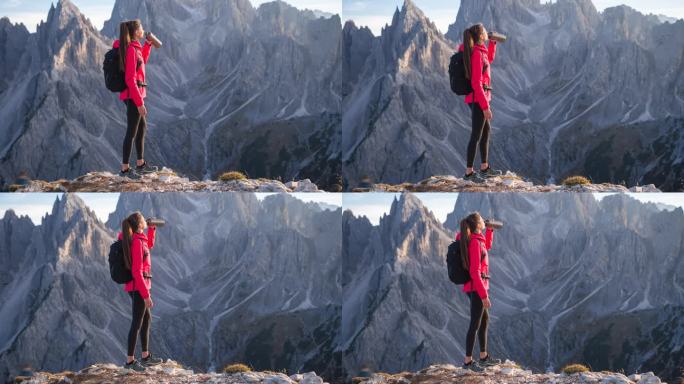 女人站在群山环绕的悬崖上，喝着水
