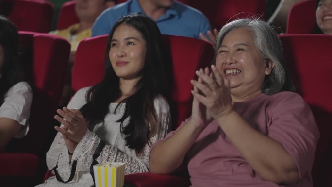 快乐的亚洲母女在影院观看喜剧电影时笑着鼓掌。一群快乐的人坐在电影院的座位上欣赏电影，鼓掌