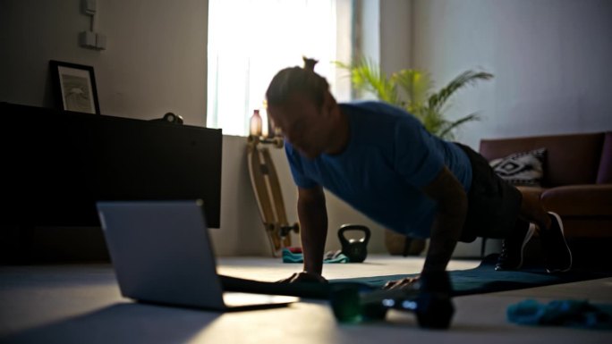 慢动作健身年轻人在客厅地板上用笔记本电脑做俯卧撑，在家做在线锻炼。力量，决心，健康的生活方式，积极的
