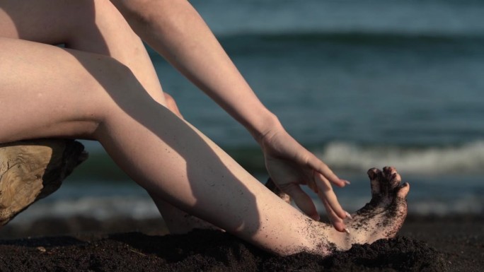 在海浪的背景下，赤脚女人在沙滩上把脚从黑沙滩上抖下来的特写