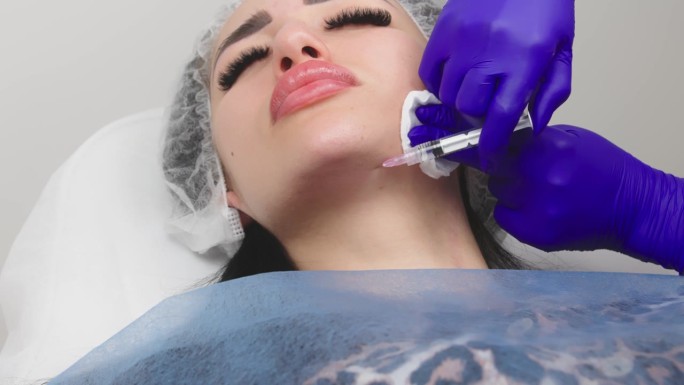 一位美容师正在给一位女士的下巴、嘴唇下面的皮肤注射含有透明质酸的生物活化剂(特写)。年轻女子在美容诊