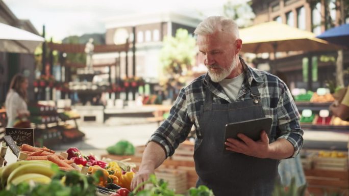 一个成年农民在市场摊位上销售新鲜有机农产品时使用平板电脑工作的肖像。中年男子在电子商务网站管理网上订