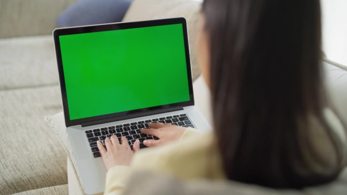 一个身份不明的女人沉浸在办公室工作的肩膀上，在沙发上舒适的家庭工作空间里使用一台带有绿屏模拟显示的笔