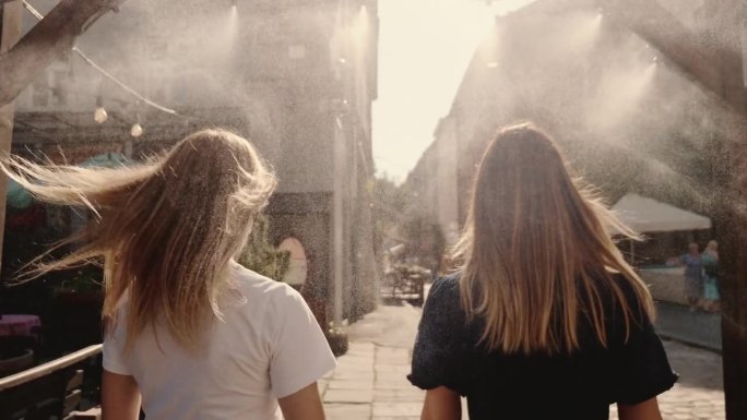 镜头跟随两个美丽快乐的年轻女性朋友，戴着圆形太阳镜走过街头喷雾，对着镜头微笑
