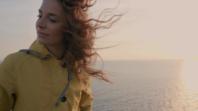 年轻女子的特写肖像与红头发吹在风中看着日落在海洋女孩穿着黄色雨衣徒步旅行在苏格兰慢动作