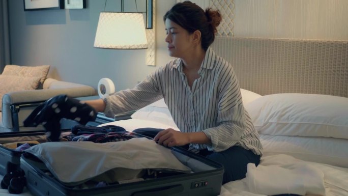 一个亚洲女人在她的酒店房间里。她的行李箱放在床上，她正在收拾衣服，因为她的旅行刚刚结束，她正要离开酒