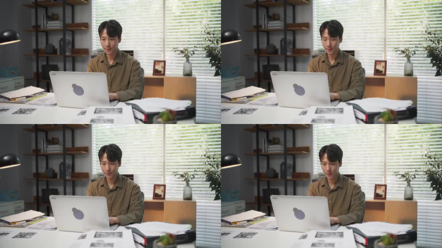 年轻英俊的韩国专业人士在他舒适时尚的办公室里坐在桌子后面的笔记本电脑上工作。亚洲专家处理文件，研究一