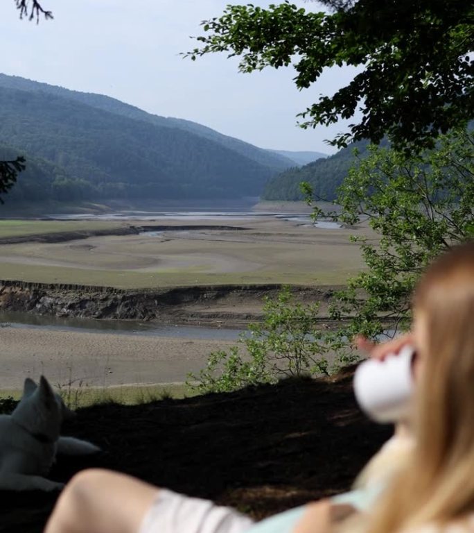 一个穿着夏装的女孩躺在山中的自然躺椅上，喝着白色马克杯里的咖啡，背景是一个干涸的湖水。垂直视频