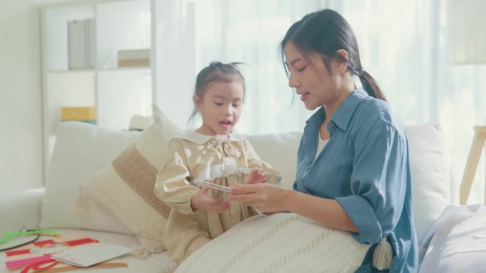 年轻的亚洲家庭妈妈和女儿在家里庆祝母亲节的时候，在客厅的沙发上互相拥抱，用心阅读手工制作的贺卡。家庭
