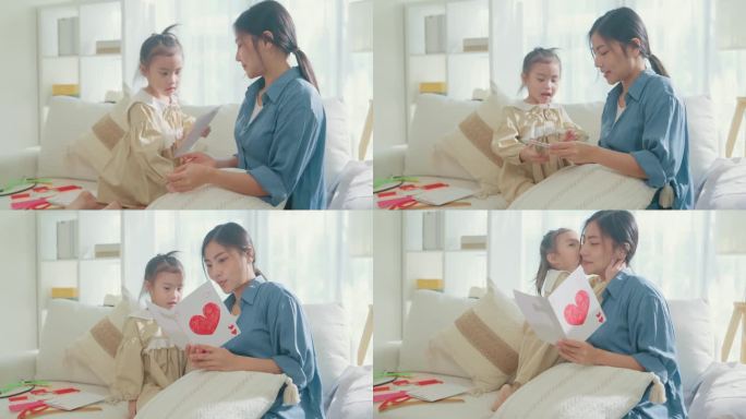 年轻的亚洲家庭妈妈和女儿在家里庆祝母亲节的时候，在客厅的沙发上互相拥抱，用心阅读手工制作的贺卡。家庭