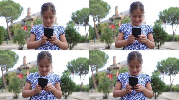 身穿蓝色印花连衣裙的黑发女孩在街上看手机。童年、电话、科技、网络、短信、快乐、放松、度假、生活理念。