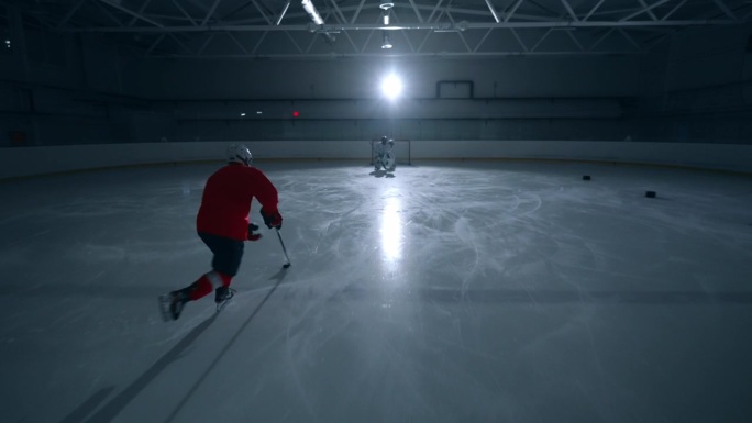 一名熟练的冰球运动员身穿红色运动衫，在黑暗的冰场上训练，快速移动并向守门员进球